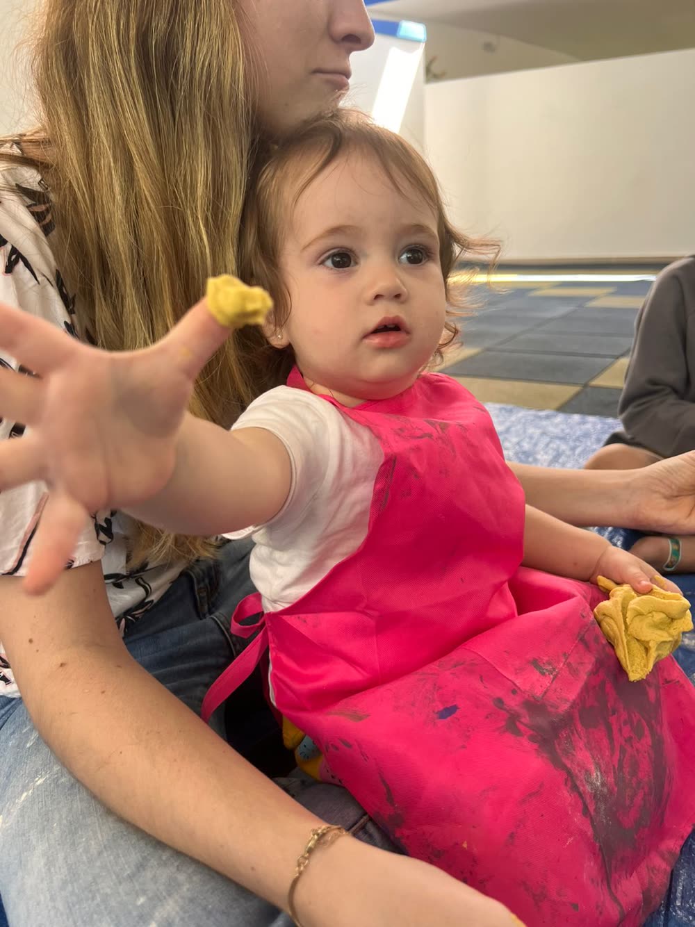 Bebê de avental rosa explorando tinta e massa com as mãos, na atividade de artes