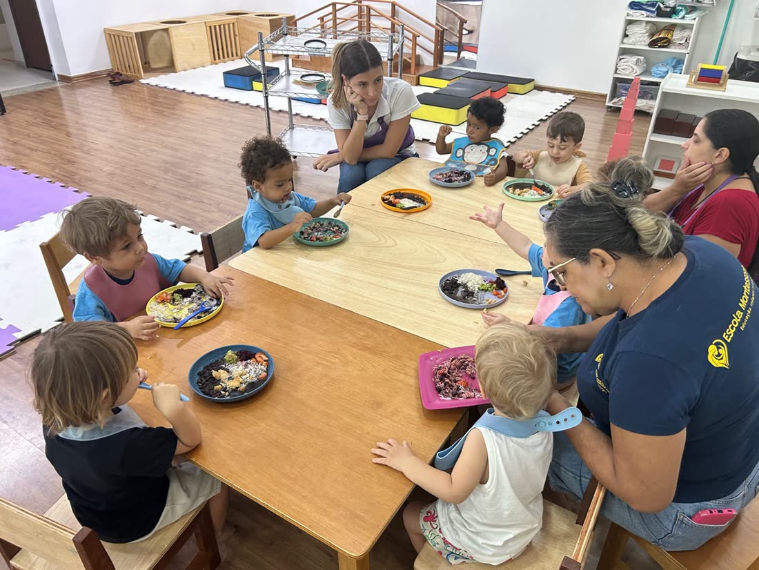 Bebês almoçando juntos à mesa baixa da creche, comendo sozinhos