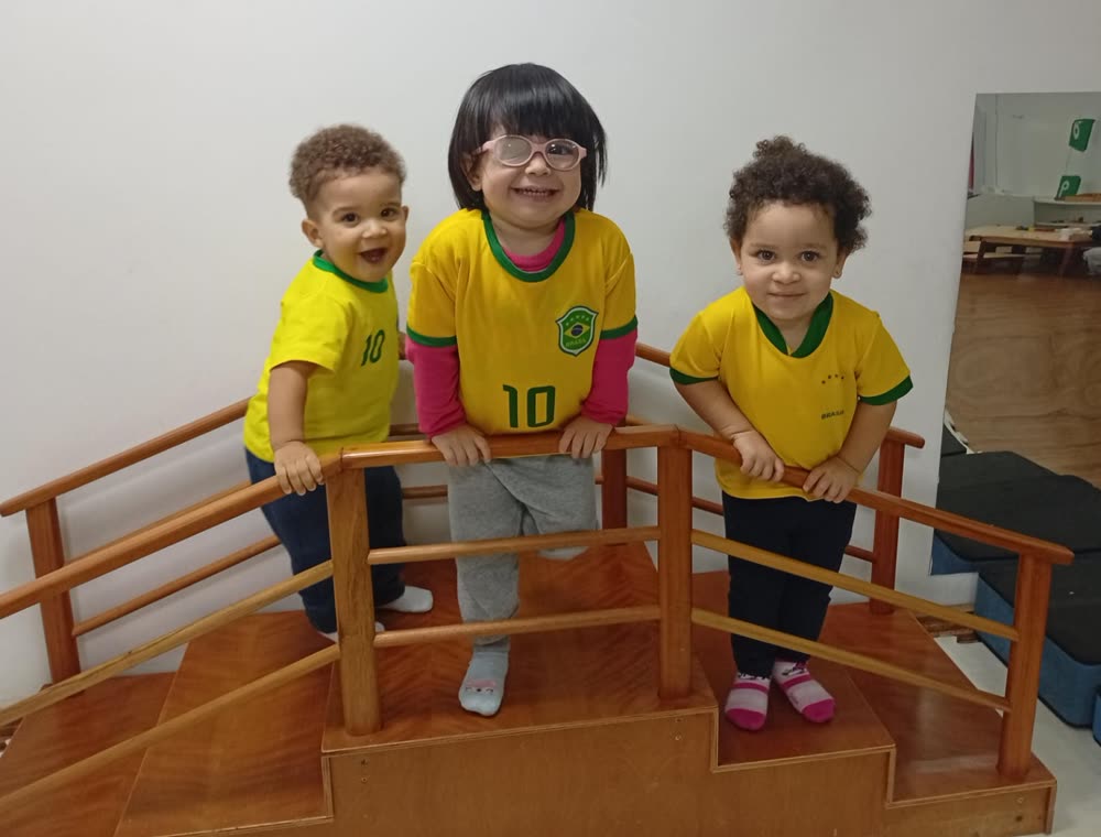 Três bebês sorrindo no alto da escada Montessori de madeira, dentro da sala