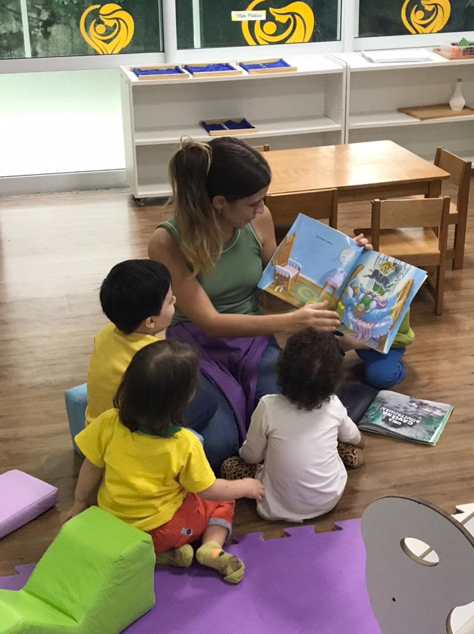 Professora lendo um livro para três bebês sentados ao redor dela