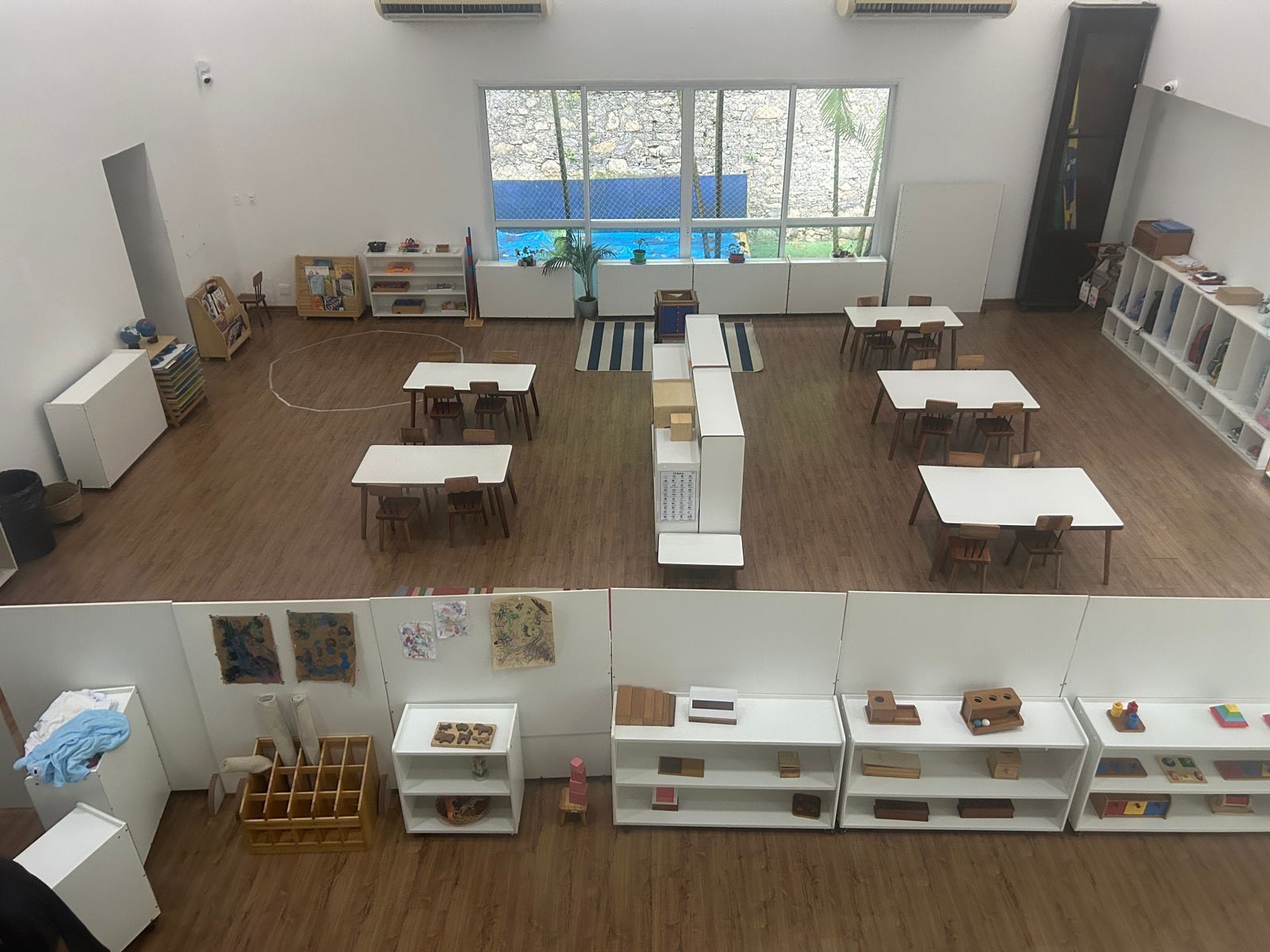 Sala ampla da Escola Montessoriana, vista de cima, com mesas baixas espalhadas, materiais ao alcance das crianças e janelas para o paredão de pedra verde