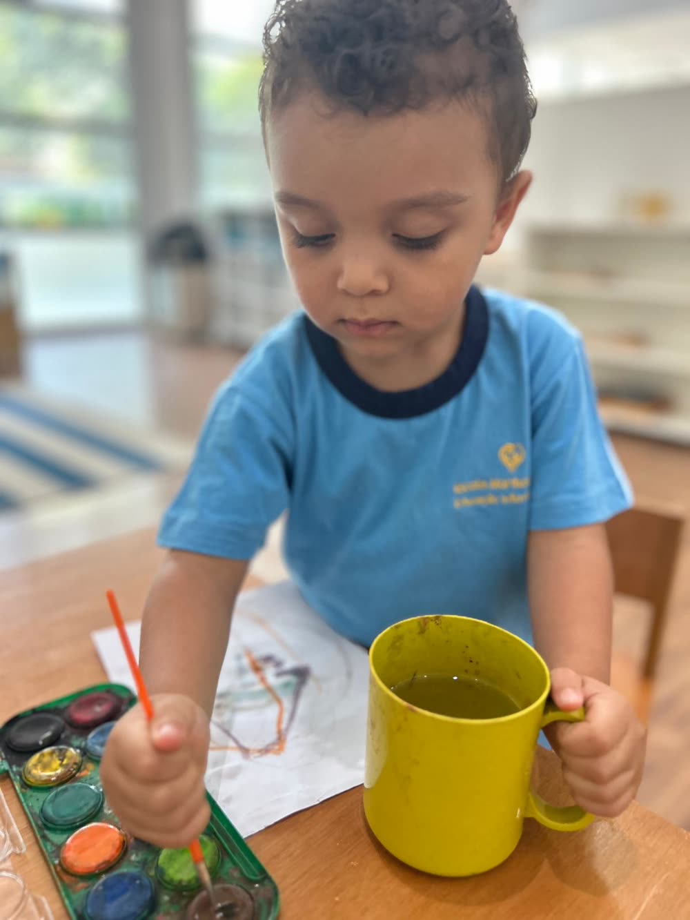 Menino concentrado pintando com pincel e aquarela na mesa da sala
