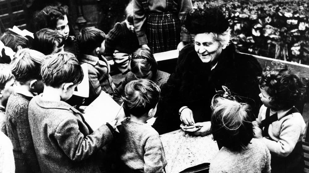 Maria Montessori idosa, sorrindo, sentada numa escola cercada de crianças que a observam de perto