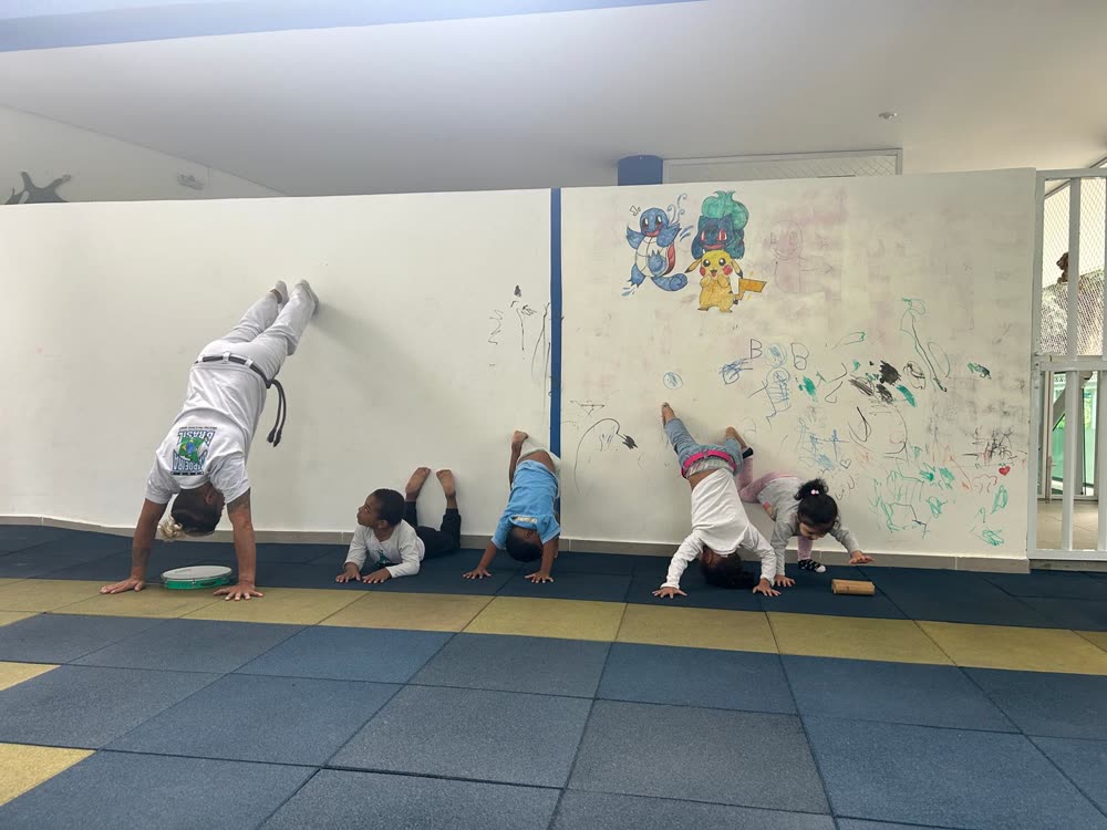 Crianças fazendo bananeira apoiadas na parede durante a aula de capoeira no pátio