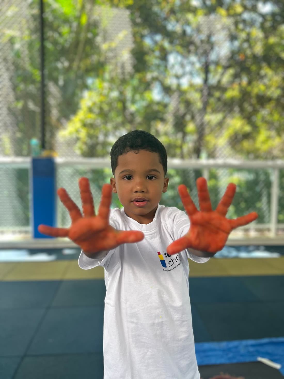 Menino mostrando as mãos pintadas, no pátio coberto, com as árvores ao fundo através da tela