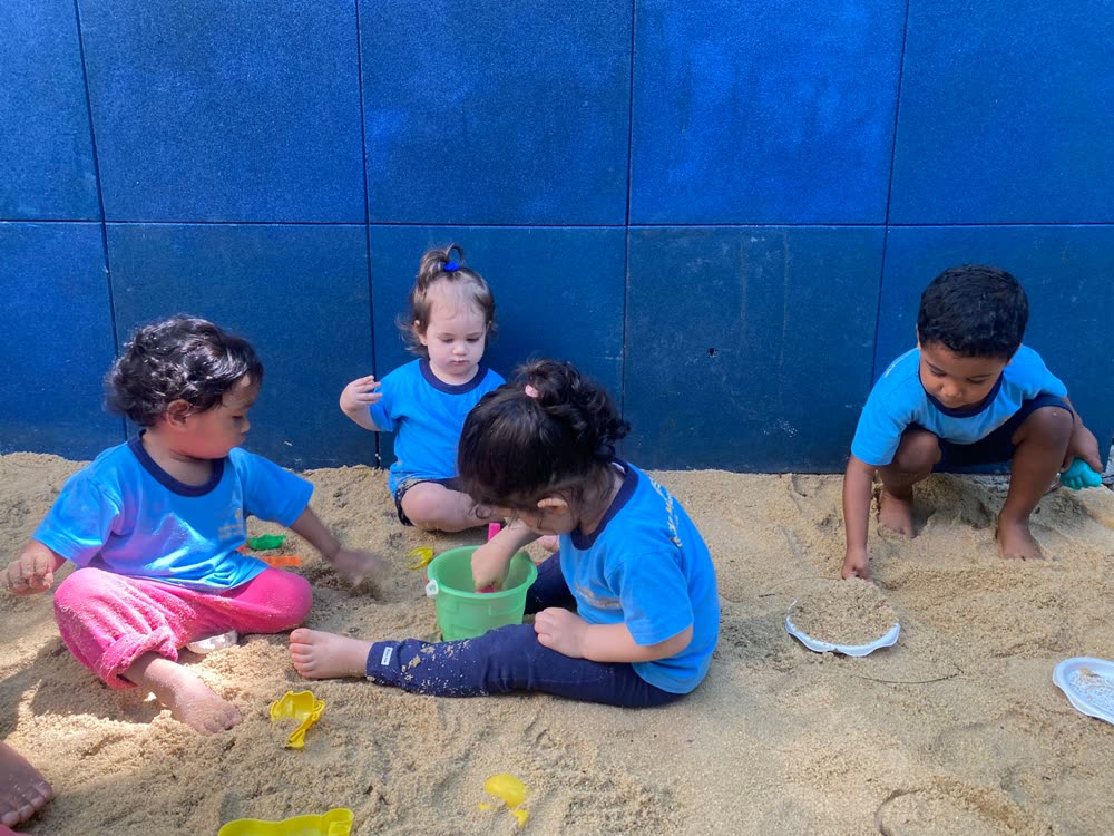 Quatro crianças de uniforme brincando juntas no tanque de areia da escola